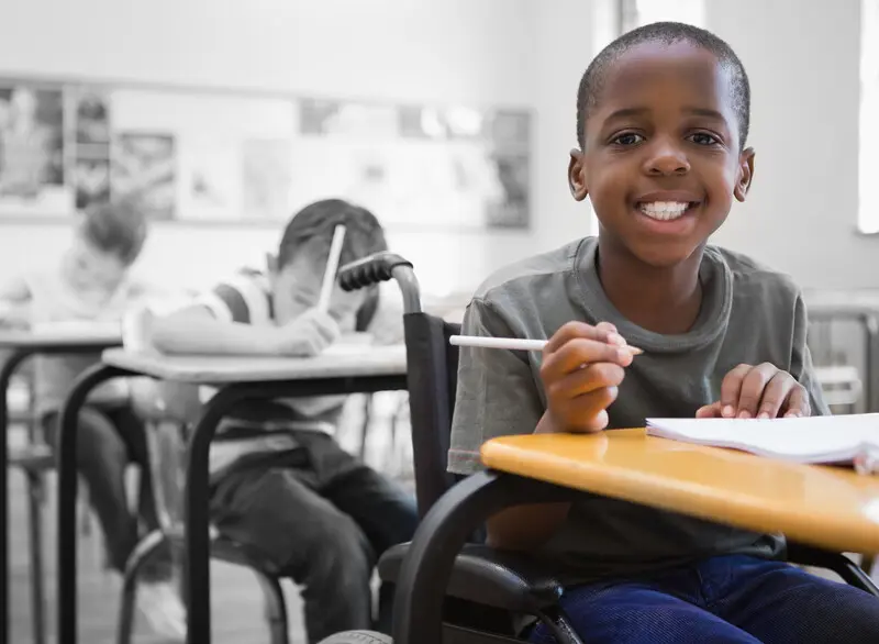 Little boy in a wheelchair, sitting at a school desk, smiling and holding a pencil