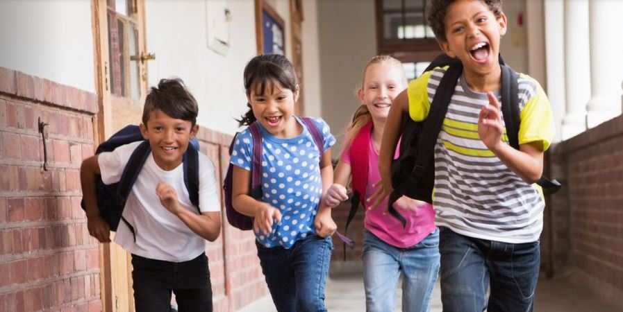 Four children with backpacks on, running down a hallway.